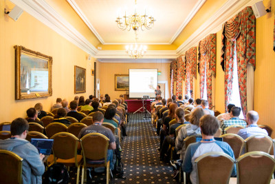 A person talking to an audience in Cardiff City Hall with a projector screen behind them a flipchart to their right.